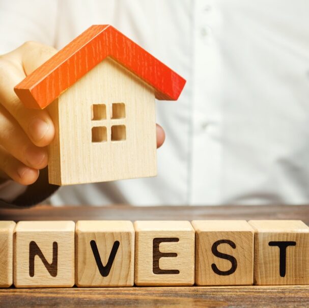 Wooden blocks with the word Invest and house in the hands of a businessman. The concept of investing in real estate construction. Effective investment. Buying apartments. Business and finance